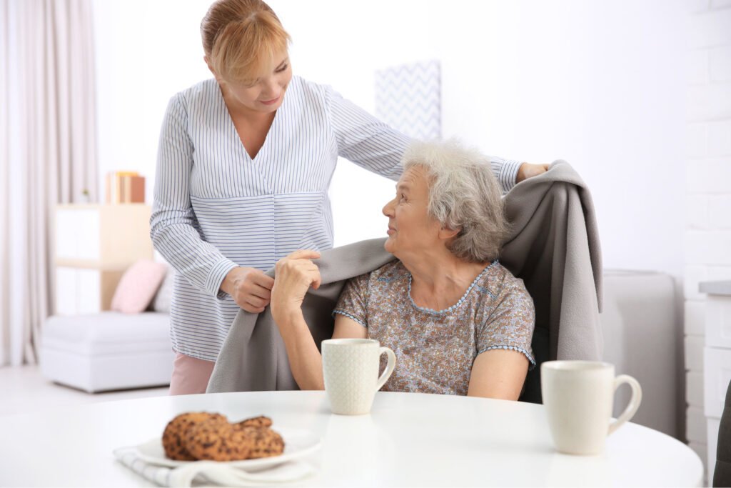 Older woman sat at table in front of mug and plate of cookies. Younger woman putting blanket over her shoulders. Atwell Care Ltd. Live in care. Home Care. Care Company. Bath. Frome. Keynsham. Saltford.