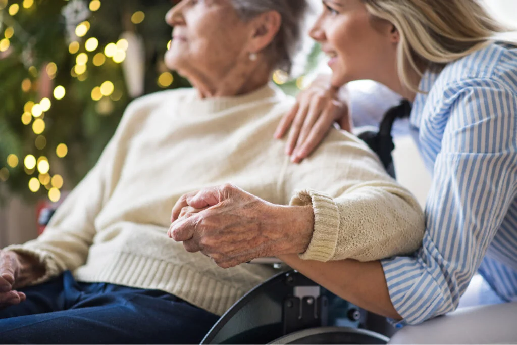 Old woman sat in wheelchair holding hands with crouched smiling younger woman. Atwell Care Ltd. Live in care. Home Care. Care Company. Bath. Frome. Keynsham. Saltford.