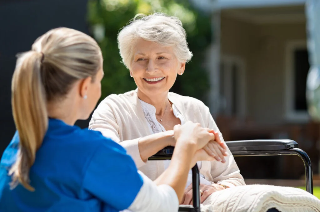 Happy old woman sat in wheelchair, hand being held by younger woman with blonde hair in ponytail. Atwell Care Ltd. Live in care. Home Care. Care Company. Bath. Frome. Keynsham. Saltford.